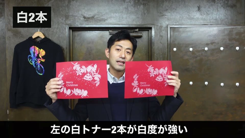 赤い色画用紙にクリスマス柄を白で印刷した2枚を並べ、左の白トナー2本のほうが白が強いことを示す様子
