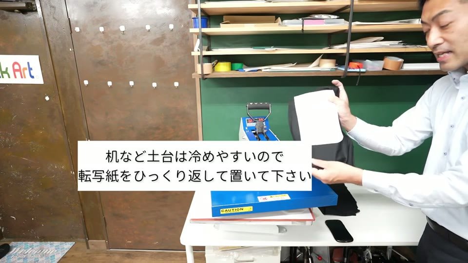 プレスしたTシャツを持ち上げて裏返している様子。テロップに「机など土台は冷めやすいので転写紙をひっくり返して置いて下さい」