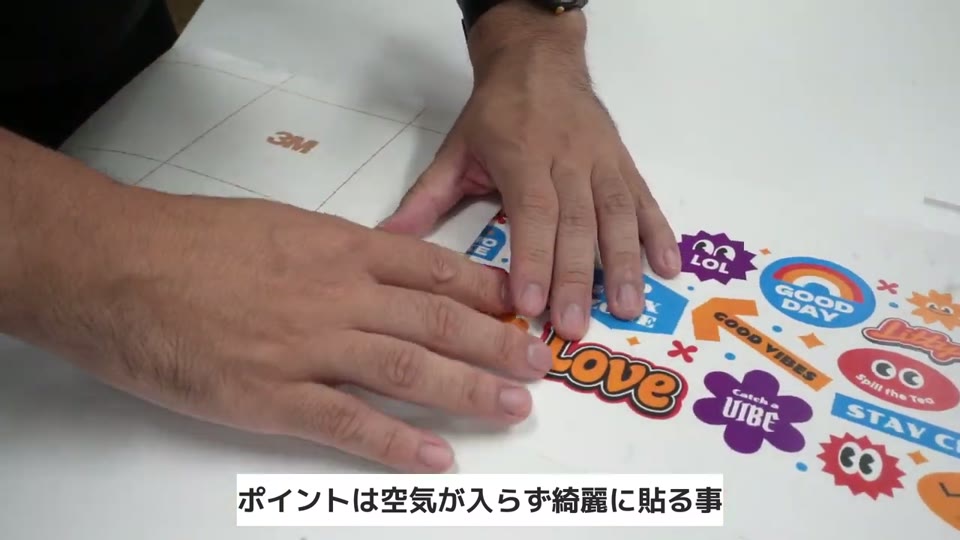 印刷したステッカーシートの上に、定規を押し付けながら保護フィルムを貼っている手元。ポイントは空気が入らず綺麗に貼る事のテロップ