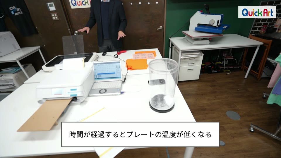 小型DTFプリンターと円筒形のパウダーケースが並ぶ作業台。時間が経過するとプレートの温度が低くなるというテロップ