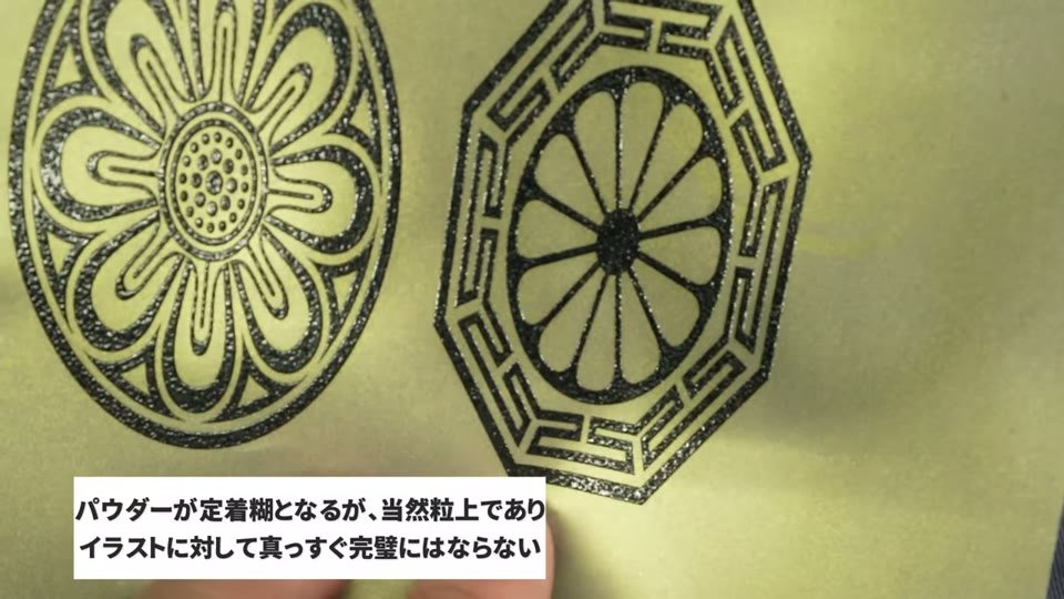 金色のDTFフィルムに黒で印刷しパウダーを付けた状態のアップ。柄の輪郭にパウダーの粒が見える