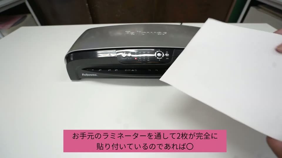 ラミネーターに通した2枚のシートを持ち上げている。テロップはお手元のラミネーターを通して2枚が完全に貼り付いているのであれば〇