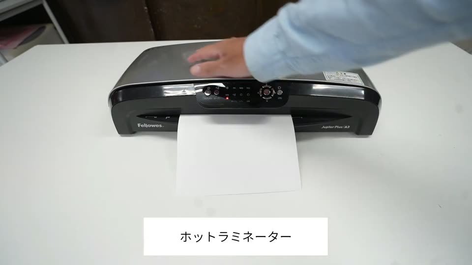 ホットラミネーターから2枚重ねのシートが出てきているところ。テロップはホットラミネーター