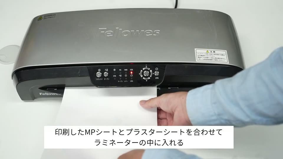 ホットラミネーターに、印刷したMPシートとプラスターシートを重ねて差し込んでいる。テロップは印刷したMPシートとプラスターシートを合わせてラミネーターの中に入れる