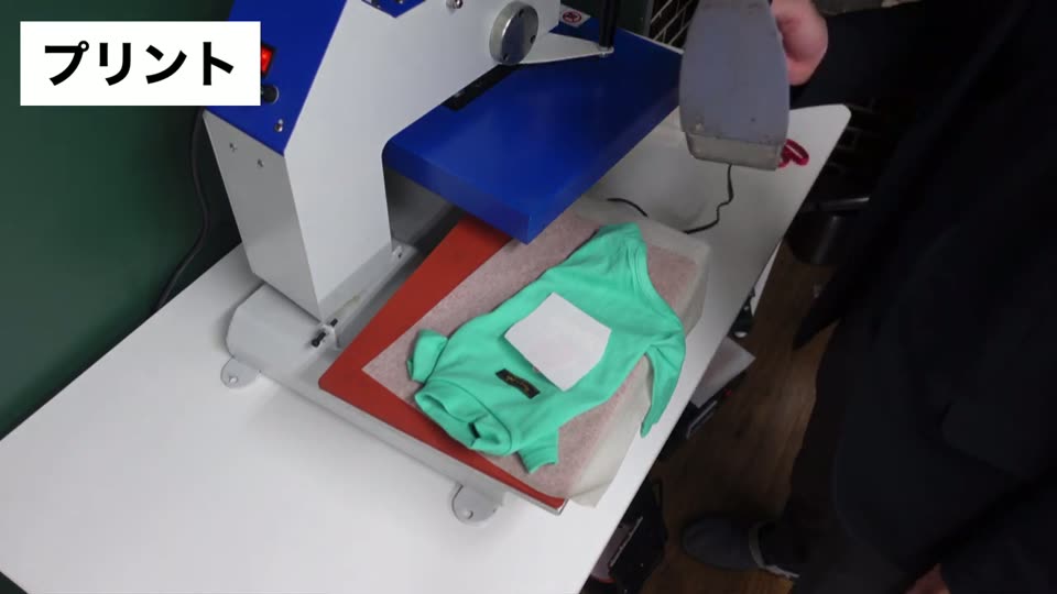 プレス機の下台に緑のペット服を広げ、マークを当てて上から家庭用アイロンで押さえている