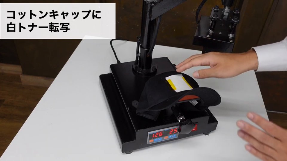 黒いコットンキャップを帽子プレス機にかぶせ、転写紙を耐熱テープで留めている。テロップ:コットンキャップも同じくマスキングが大事です