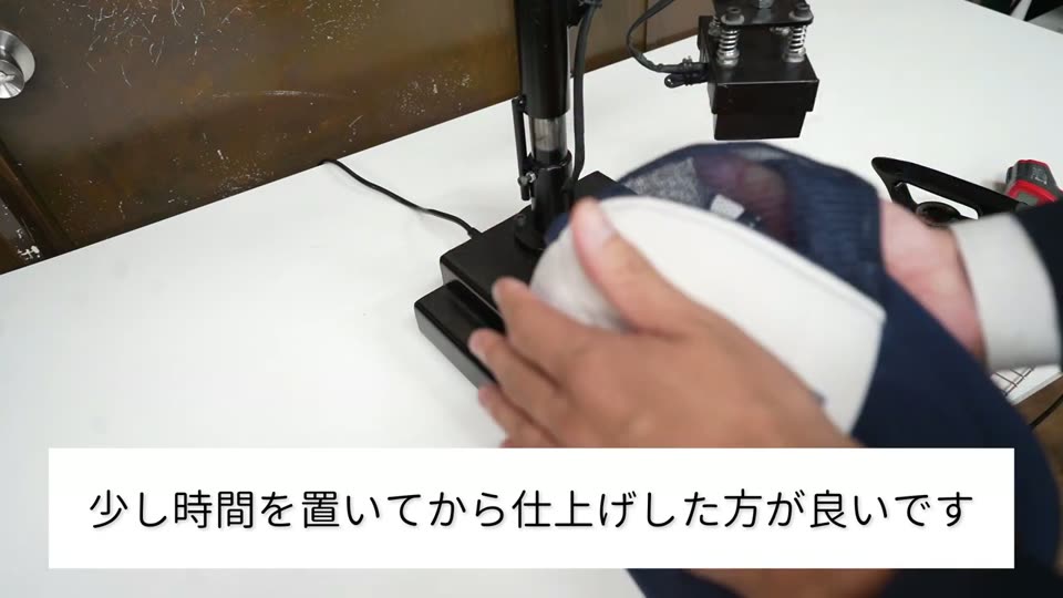 帽子プレス機の横でメッシュキャップを手に取っている様子と、少し時間を置いてから仕上げした方が良いですというテロップ