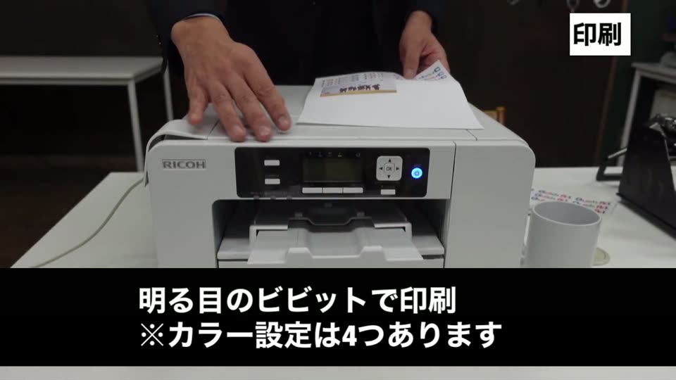 リコー製の昇華プリンターから転写紙が出てくる様子。明るめのビビッドで印刷、カラー設定は4つと説明するテロップ