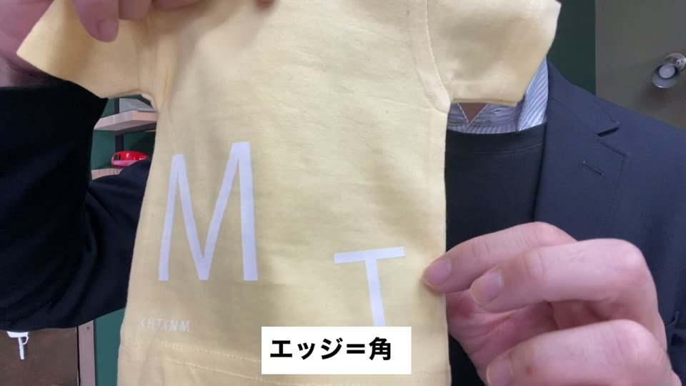 黄色いミニTシャツにプリントした白いMとTの文字の角を指さし、エッジ=角と字幕が出ている様子