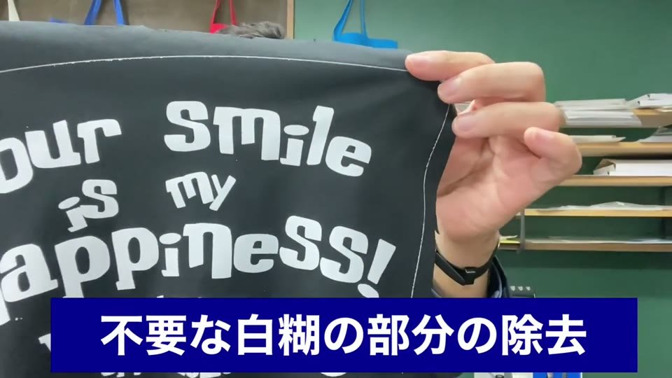 黒い布に白い文字をプリントしたもので、4辺を切らずに転写したため周囲に白い糊の枠がついている様子と、不要な白糊の部分の除去と字幕が出ている様子