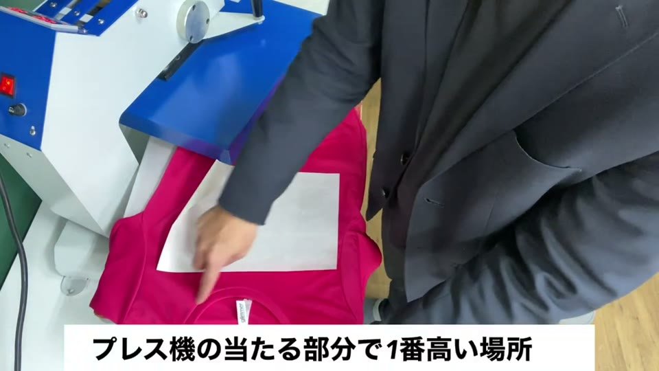 ピンクのTシャツをプレス機の下ゴテに着せ、襟ぐりの位置を指さしながらプレス機の当たる部分で1番高い場所と字幕が出ている様子