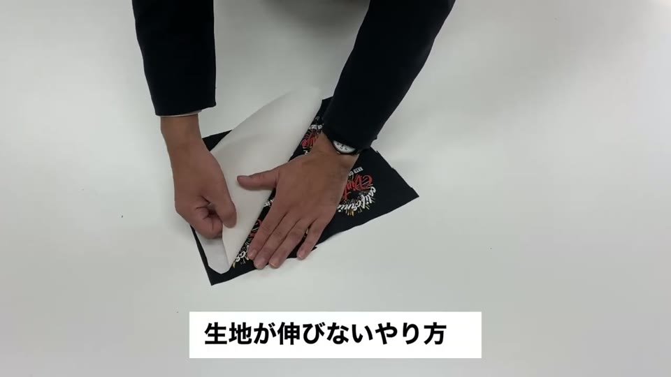 右手で転写紙を少し持ち上げ、左手で黒い生地を押さえながら剥がしている手元と、生地が伸びないやり方と字幕が出ている様子