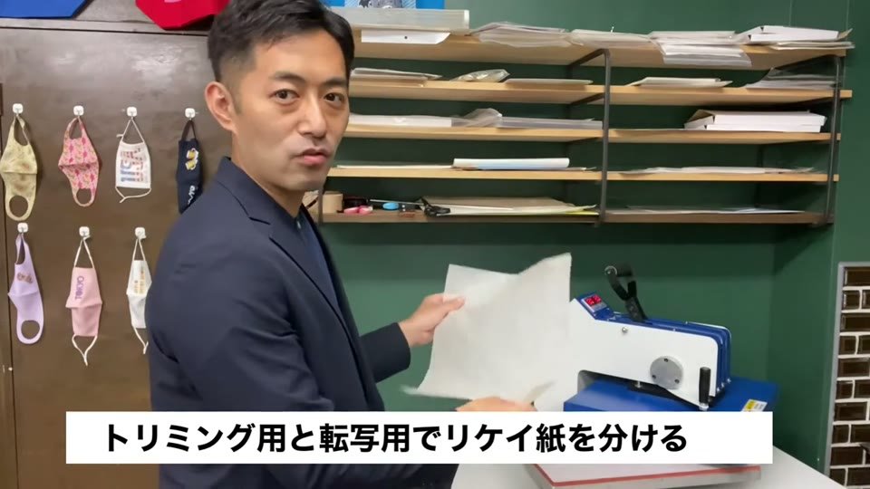 プレス機の横でリケイ紙を手に持ち、トリミング用と転写用でリケイ紙を分けると字幕が出ている様子