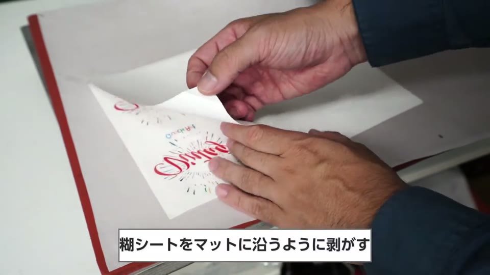 プレス機の下ゴテの上で、赤い文字を印刷したMP93シートから糊シートを低い角度で剥がしている手元
