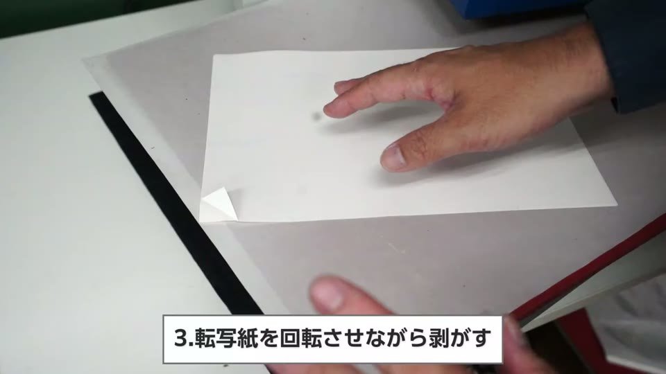 プレス台の上で用紙を少し回転させながら糊シートを剥がしている手元と、転写紙を回転させながら剥がすの字幕