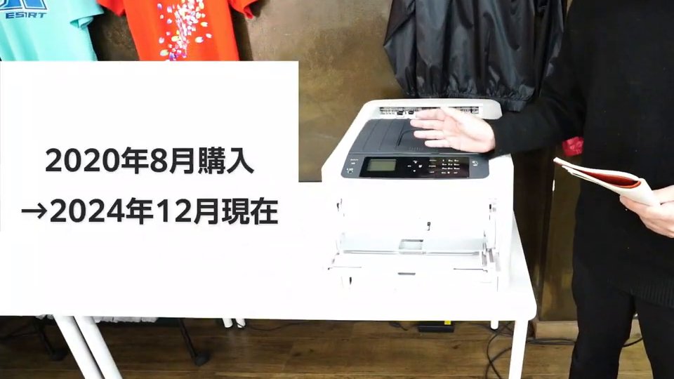 ショールームの机に置かれた白いレーザープリンター。テロップは「2020年8月購入→2024年12月現在」